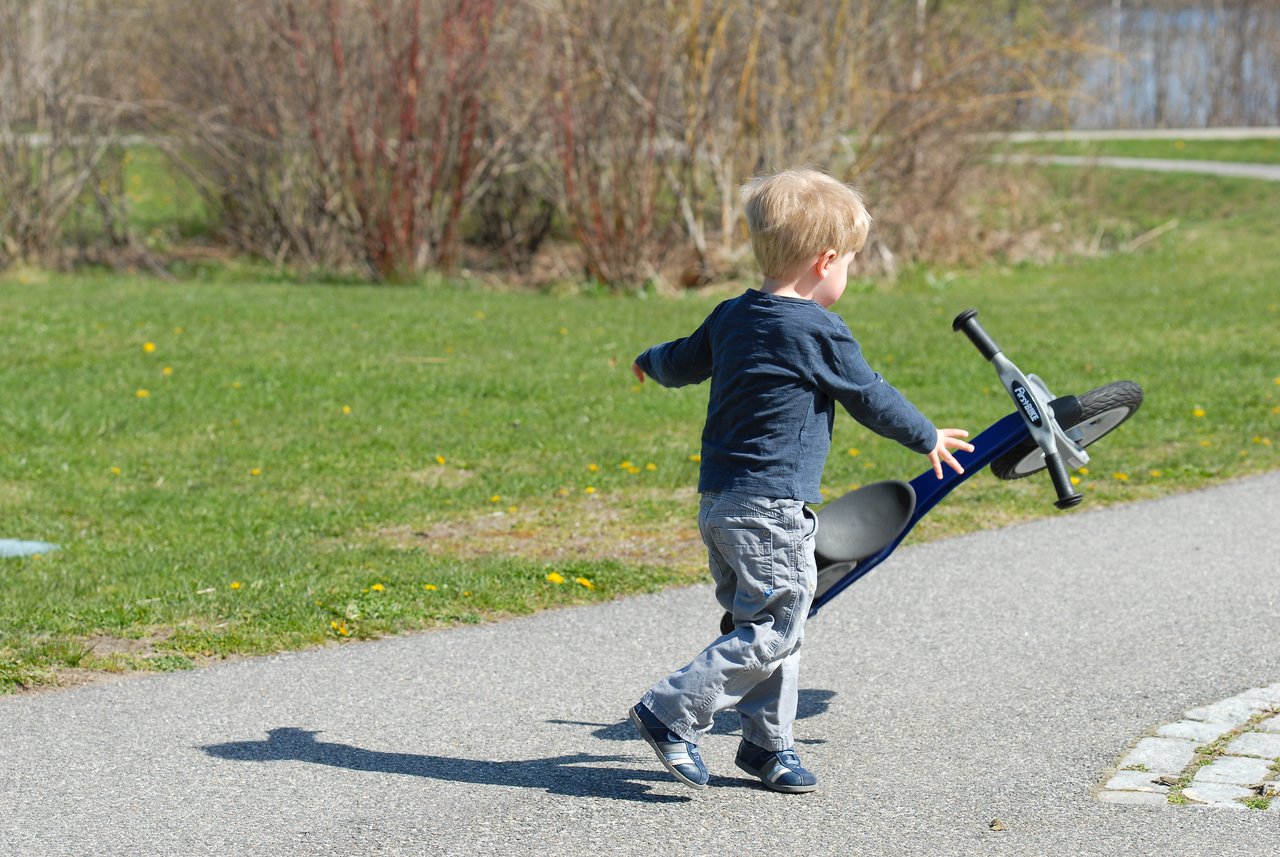 A young child in a blue shirt throws a small bike onto a paved path in a grassy park.
