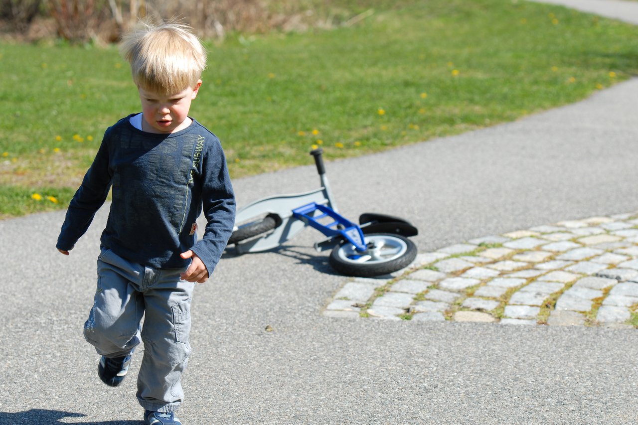 A young child walks away from a fallen blue balance bike on a paved path in a park.