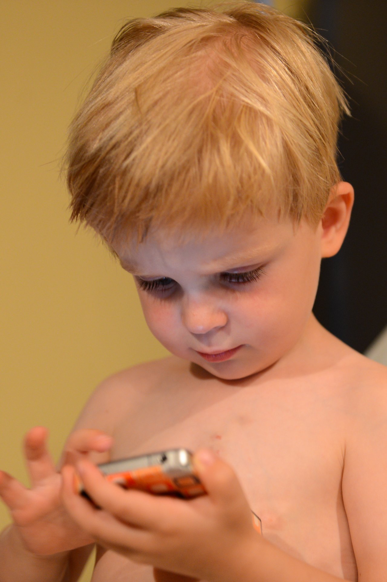 A young child with blond hair looks at a smartphone, tapping the screen with both hands.