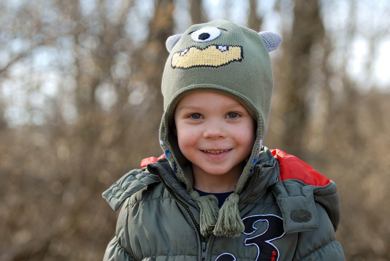 A young child wearing a green monster hat and winter coat smiles while standing outdoors in a wooded area.