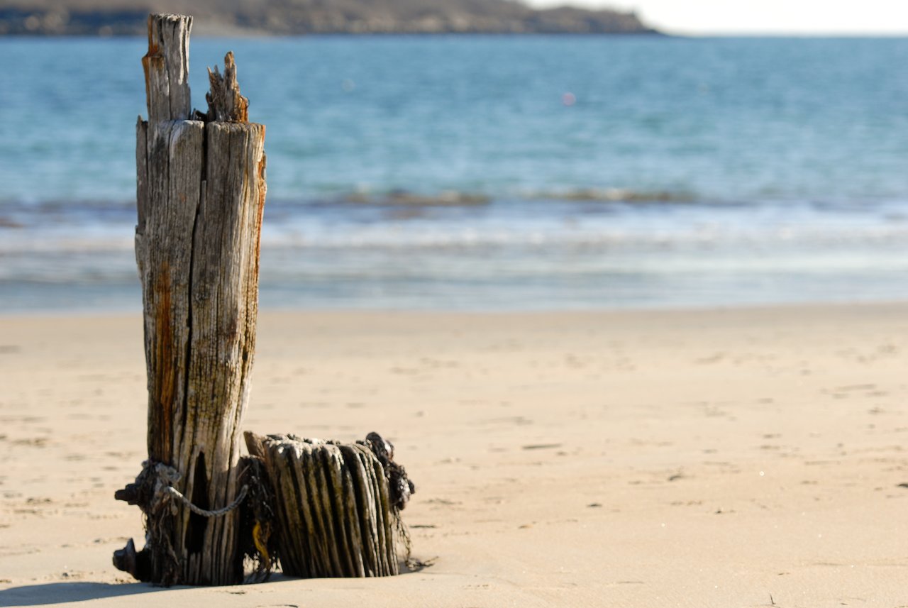 A weathered wooden post stands in the sand near the shoreline, with the ocean and distant land in the background.
