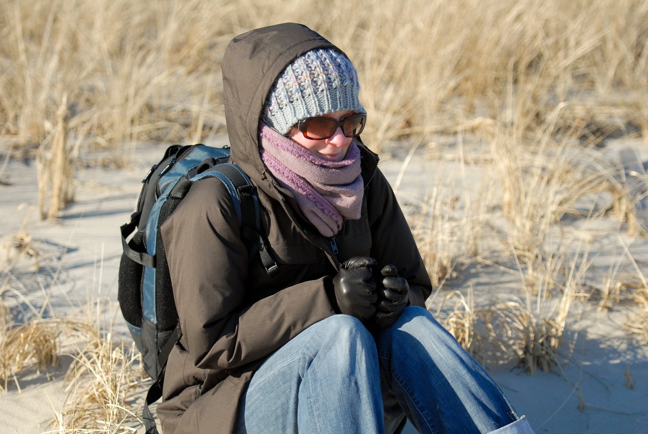 A person in warm clothing sits on a sandy beach, bundled up with gloves and a scarf on a cold day.