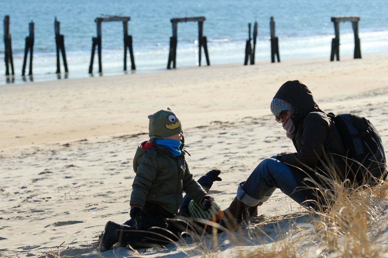 A child and an adult sit on a sandy beach, dressed in warm clothing, facing each other.