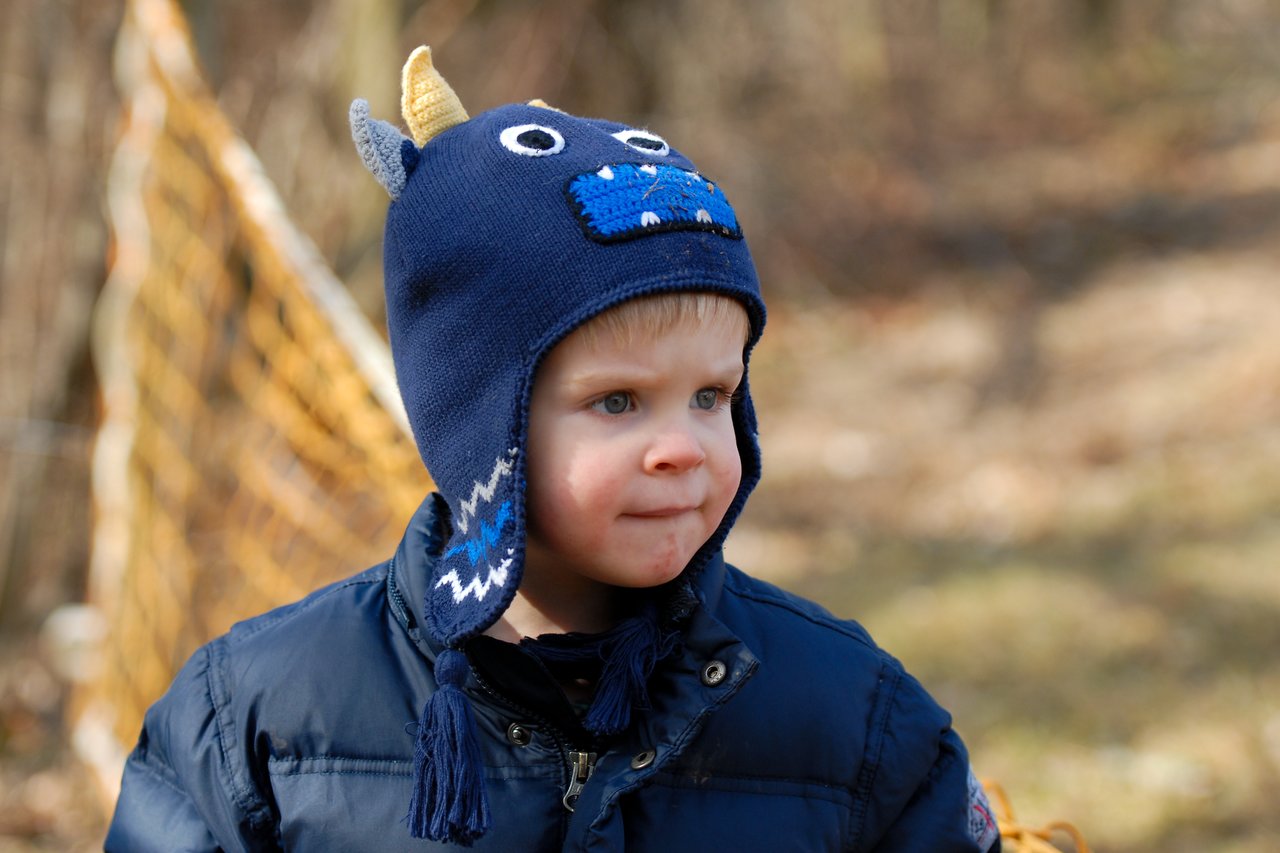 A young child in a blue monster hat and winter coat looks off to the side outdoors.