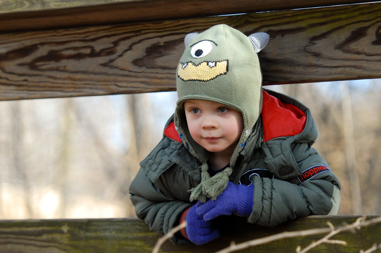 A young child in a green monster hat and gloves leans on a wooden fence, looking ahead with curiosity.