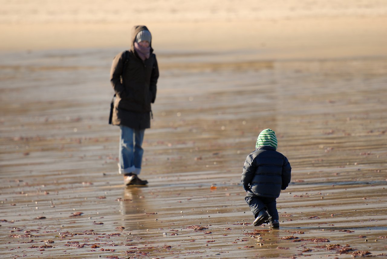 A child in a hooded jacket walks toward an adult on a wet, sandy beach.