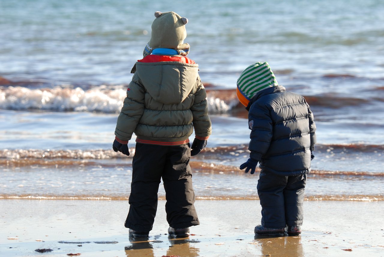 Two children in winter clothing stand on a wet sandy beach, looking at the water and shoreline.
