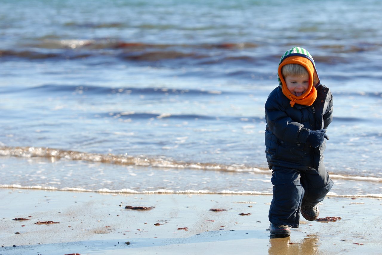 A young child in a winter coat walks along the wet sand near the water's edge at the beach.