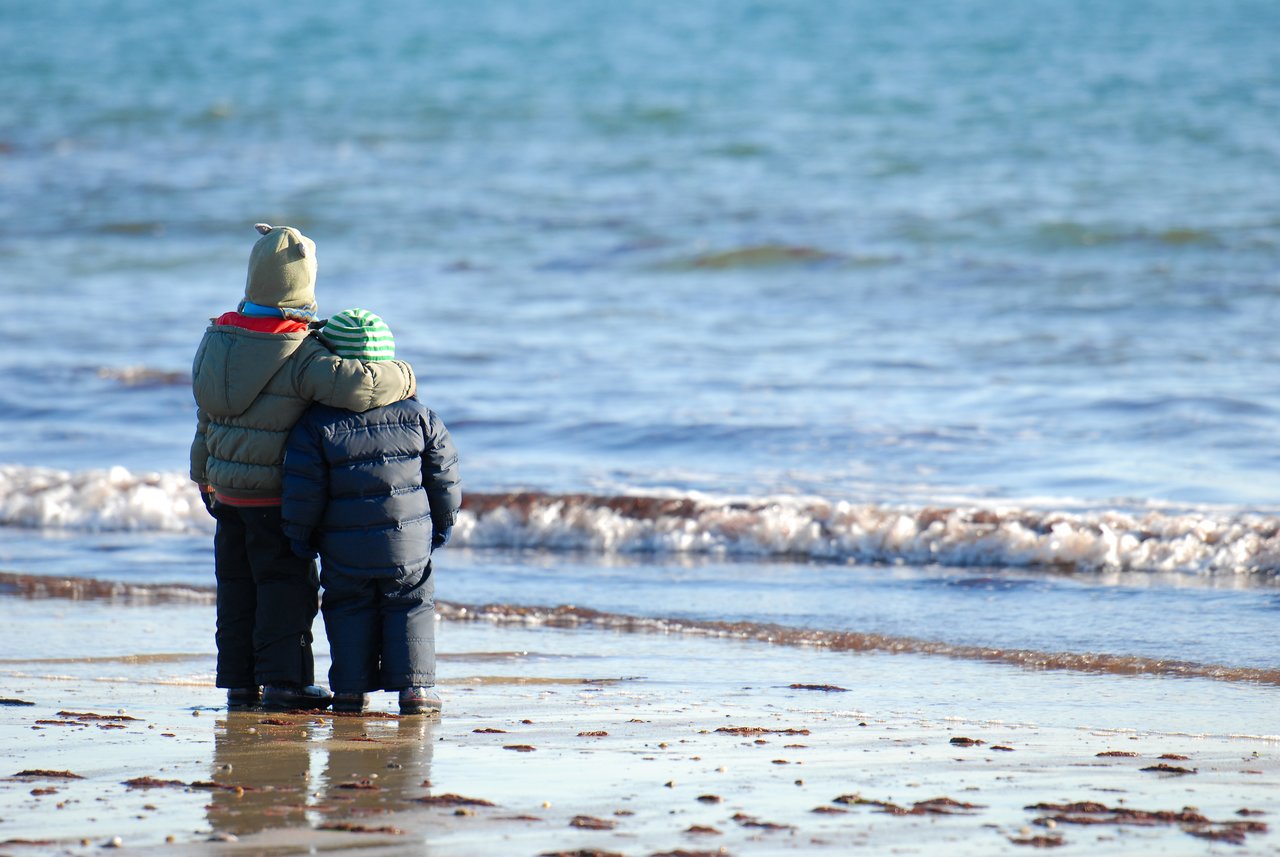 Two children in winter coats stand on a wet beach, facing the ocean, with one arm around the other.