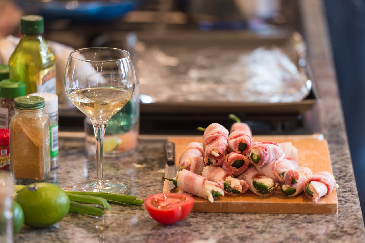 A cutting board with bacon-wrapped jalapeño poppers, surrounded by ingredients and a glass of white wine.