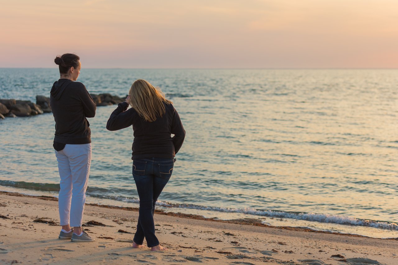Two people stand on a sandy beach, facing the ocean, wearing jackets.