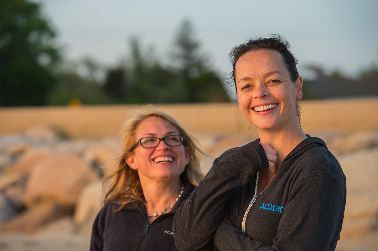 Two people stand outdoors, smiling and laughing while wearing matching dark jackets.