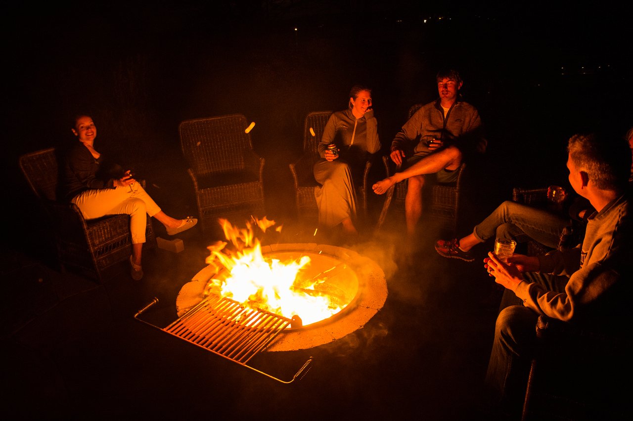A group of friends sits around a campfire at night, talking and holding drinks.