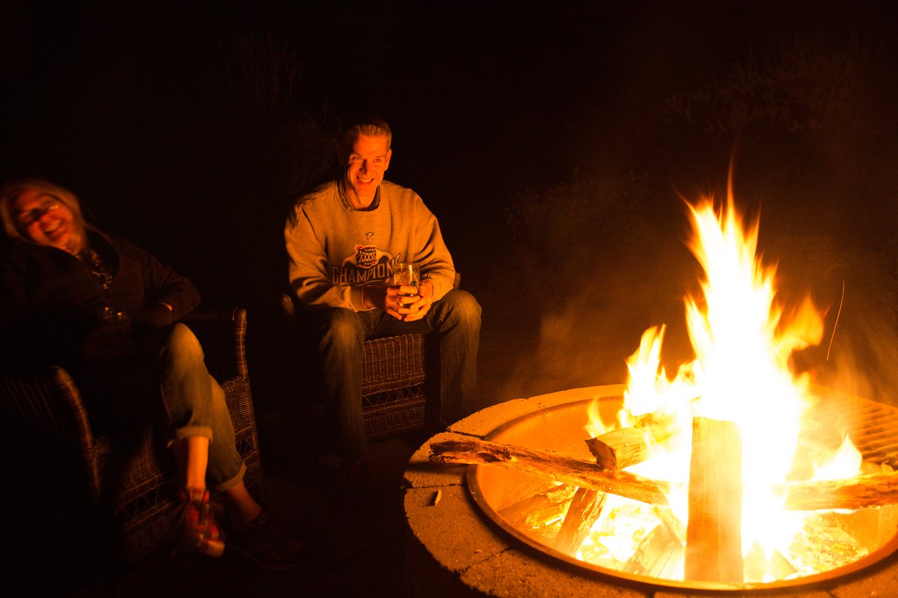 Two friends sit around a campfire at night, smiling and enjoying the warmth.