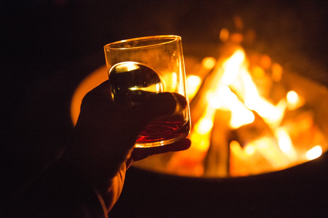 A person holds a glass with a drink near a campfire at night.