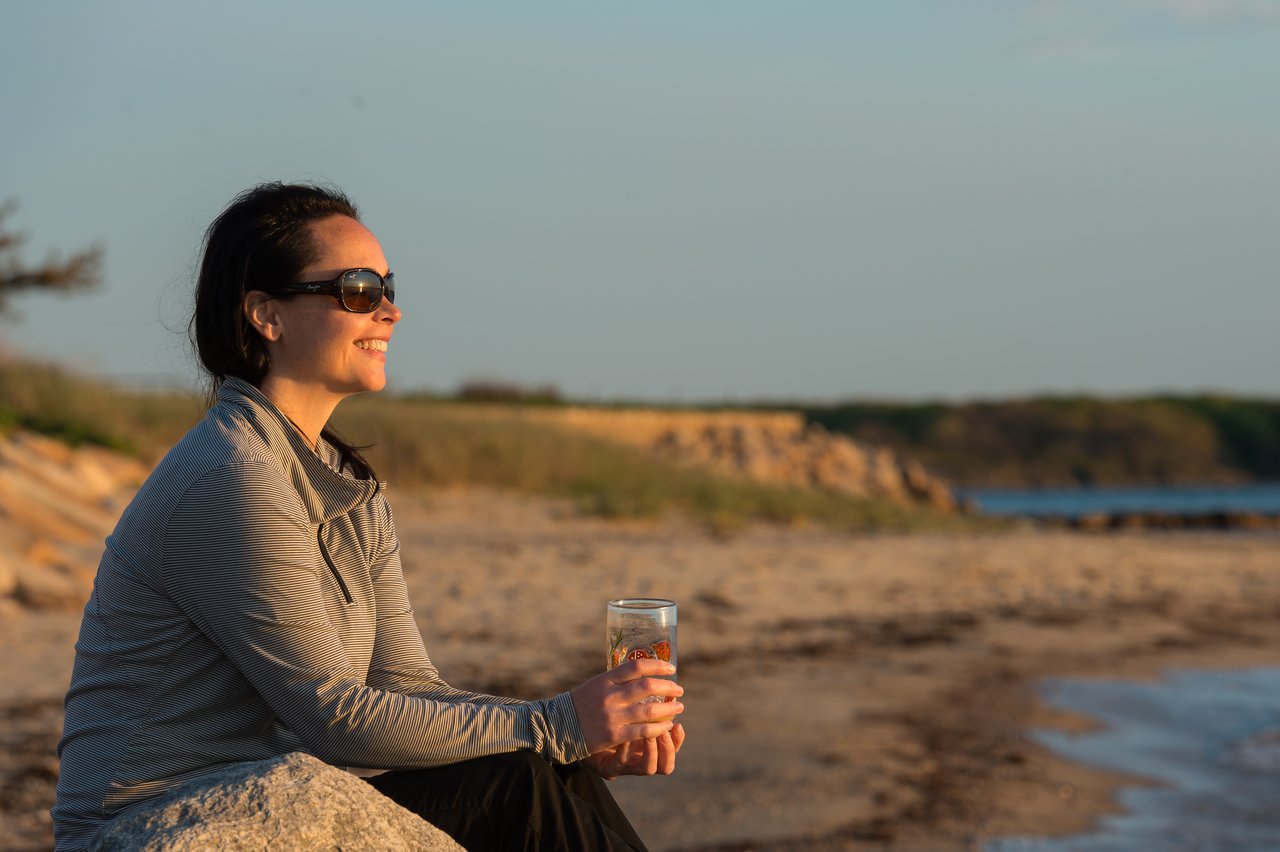 A person wearing sunglasses sits on a beach, holding a glass of gin and tonic while smiling.