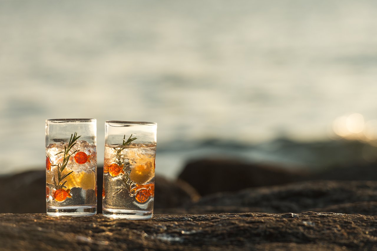 Two glasses of gin and tonic with ice, fruit, and rosemary sit on a rock near the water.