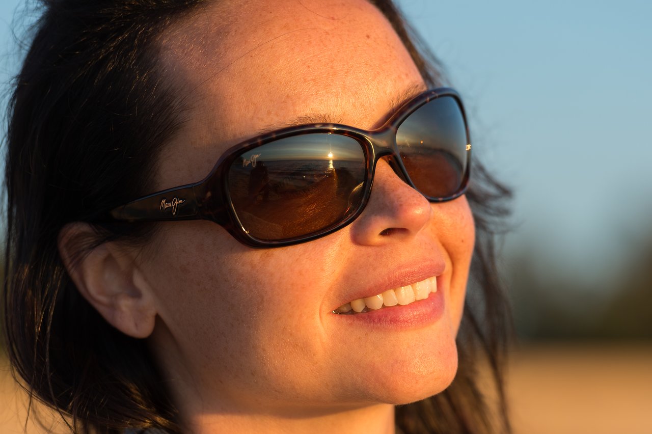 A person wearing sunglasses smiles, with a beach and sunset reflected in the lenses.