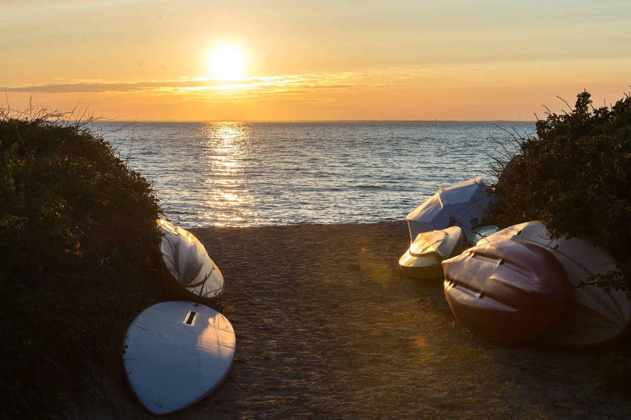 Several small boats rest on a sandy beach at sunset, with the ocean and sun in the background.