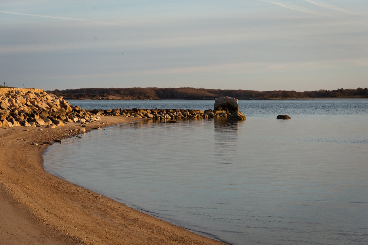 A calm shoreline with a rocky barrier extending into the water, surrounded by still water and distant trees.