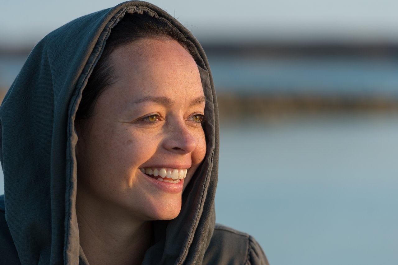 A person wearing a hooded jacket smiles while looking off to the side near a calm body of water.
