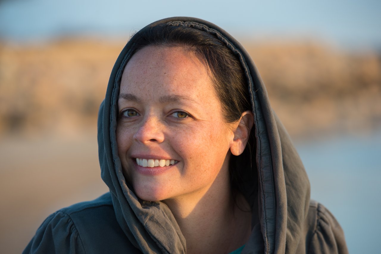 A woman wearing a hooded jacket smiles at the camera with a blurred outdoor background.