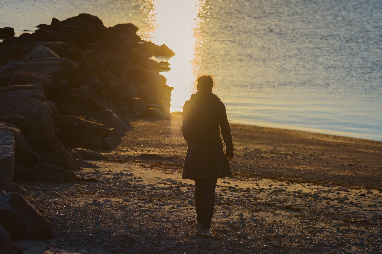 A person in a coat walks along a rocky shoreline toward the water, with sunlight reflecting on the surface.