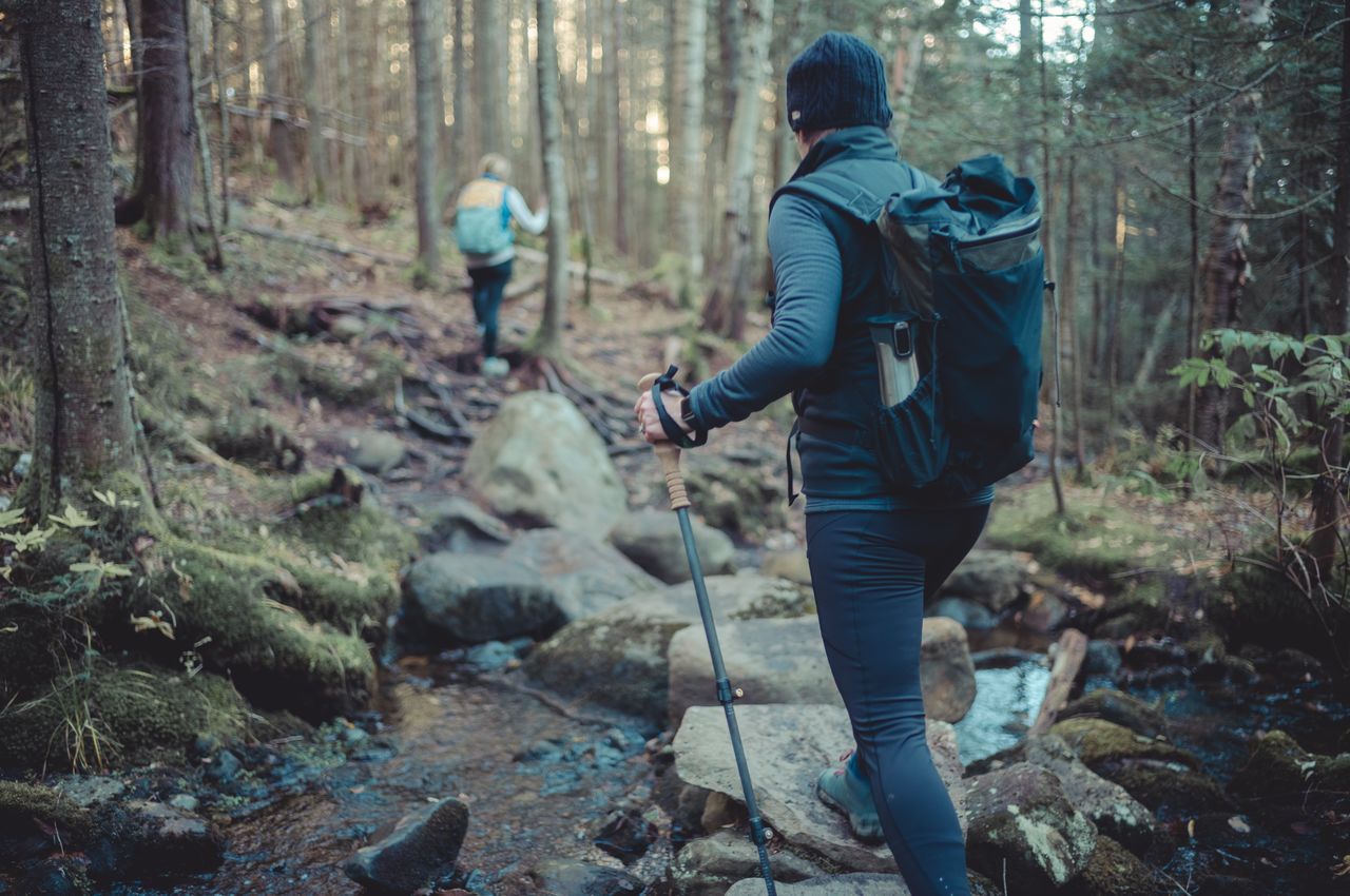 A woman hikes through the woods, crossing a stream by stepping on rocks and using trekking poles to keep her balance.