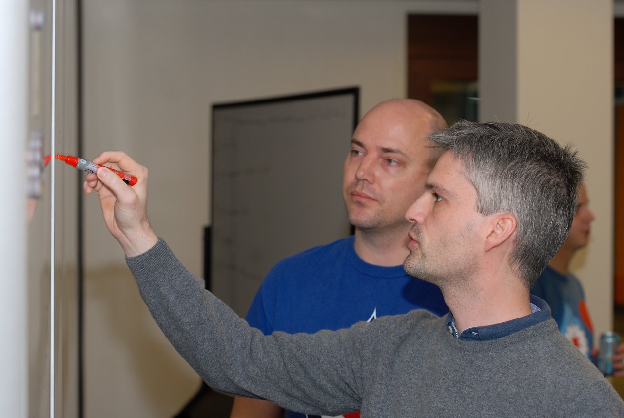 A man writes on a whiteboard with a red marker while another person observes closely.