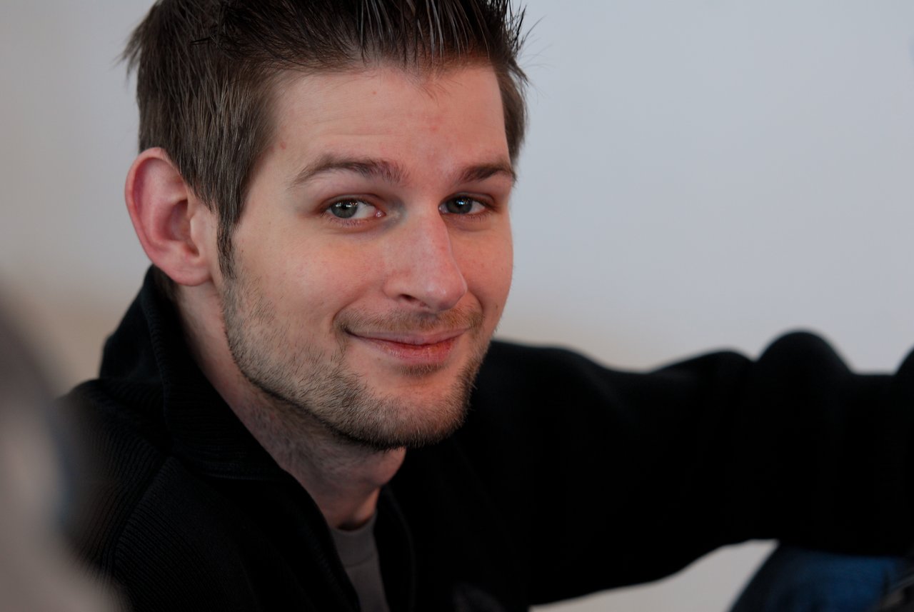 A man with short hair and a beard smiles slightly while sitting indoors, looking towards the camera.