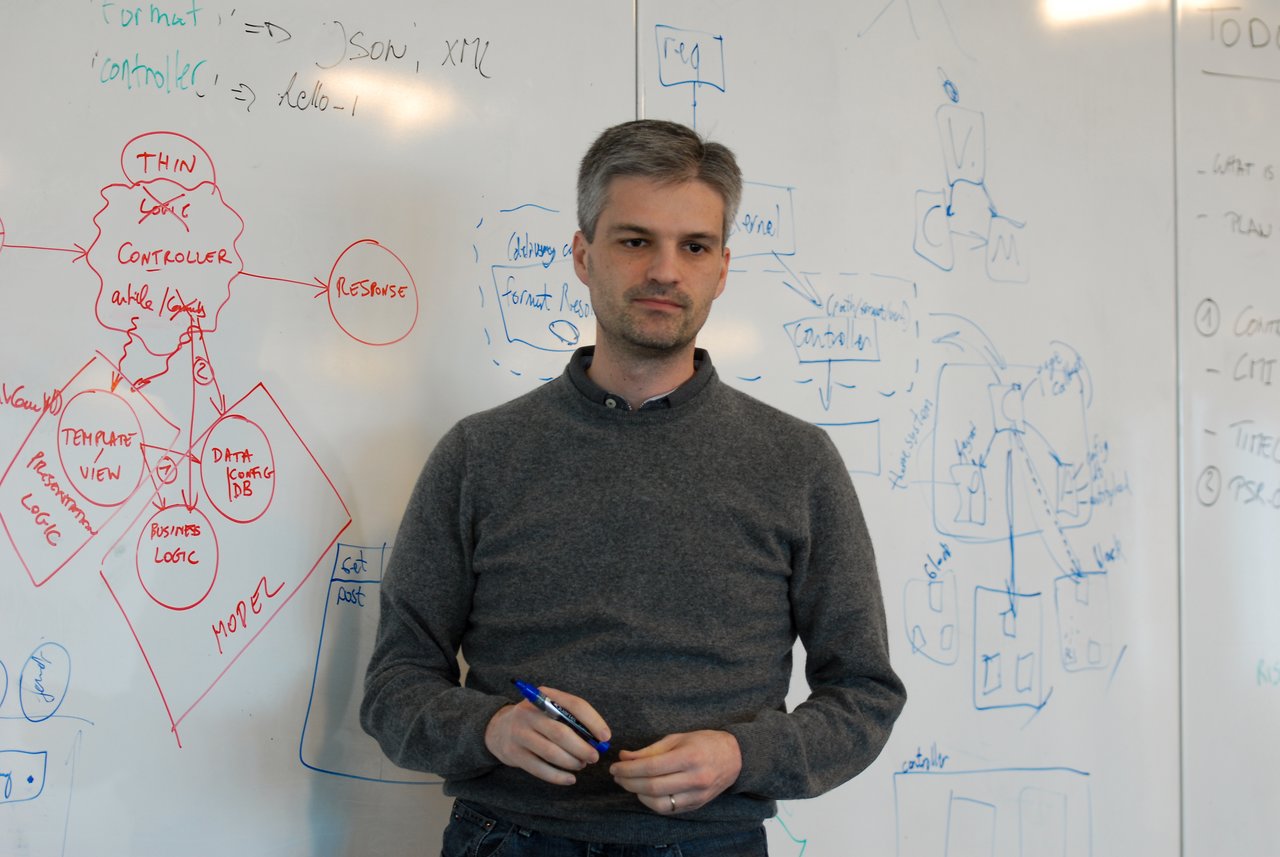 A man stands in front of a whiteboard filled with diagrams, holding a pen during a discussion or presentation.