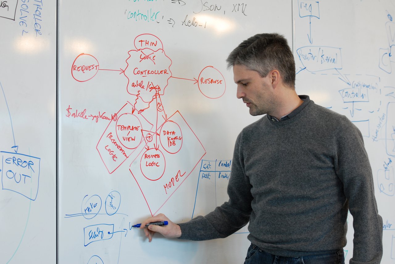 A man in a gray sweater explains a diagram on a whiteboard while pointing with a blue marker.