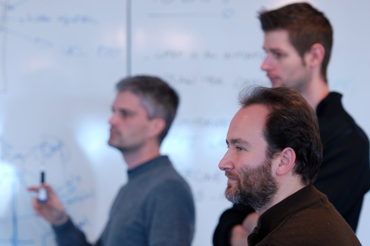 Three men in a discussion, one writing on a whiteboard while the others observe attentively.