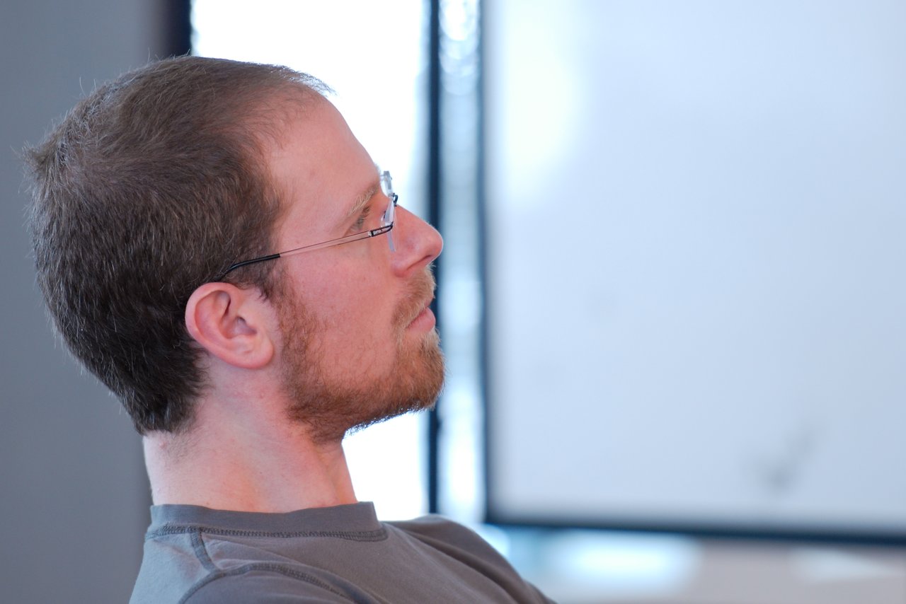 A person with glasses and a beard sits attentively, looking forward.
