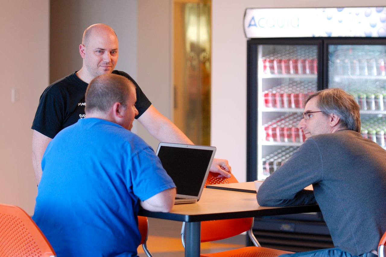 Three people collaborate at a table, with one using a laptop and another standing while speaking.