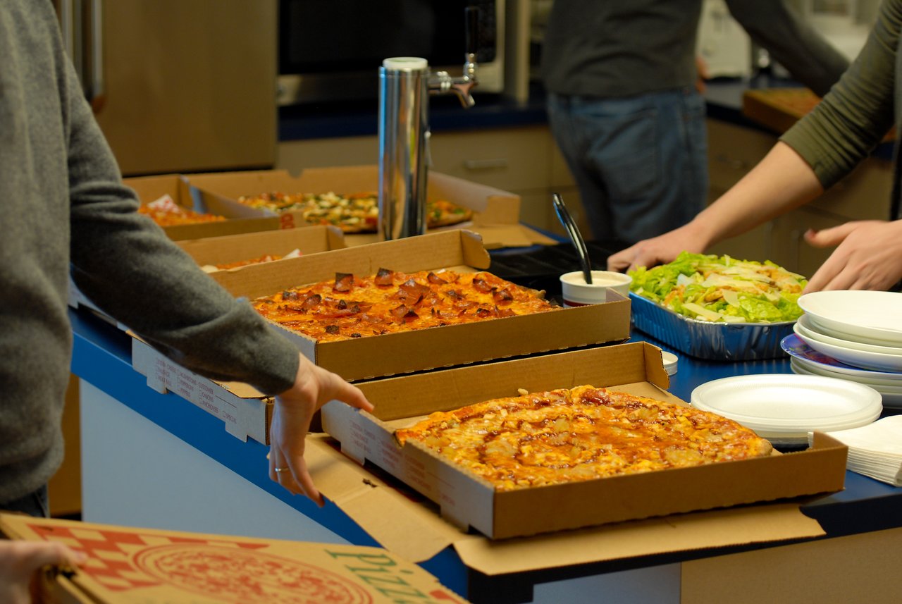 Several people serve themselves pizza and salad from an open buffet in a kitchen or break room.