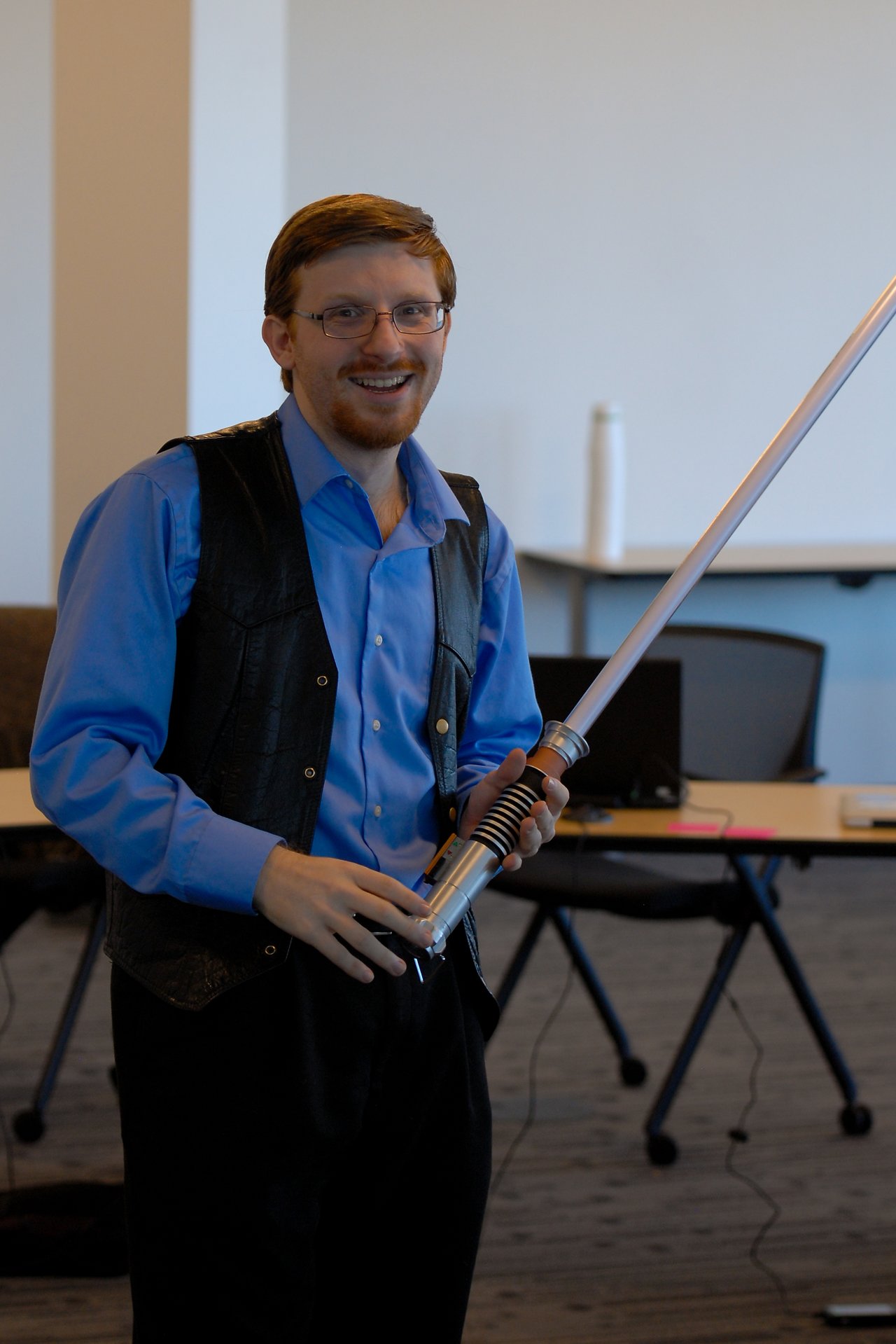 A smiling person in a blue shirt and vest holds a lightsaber in an office setting.
