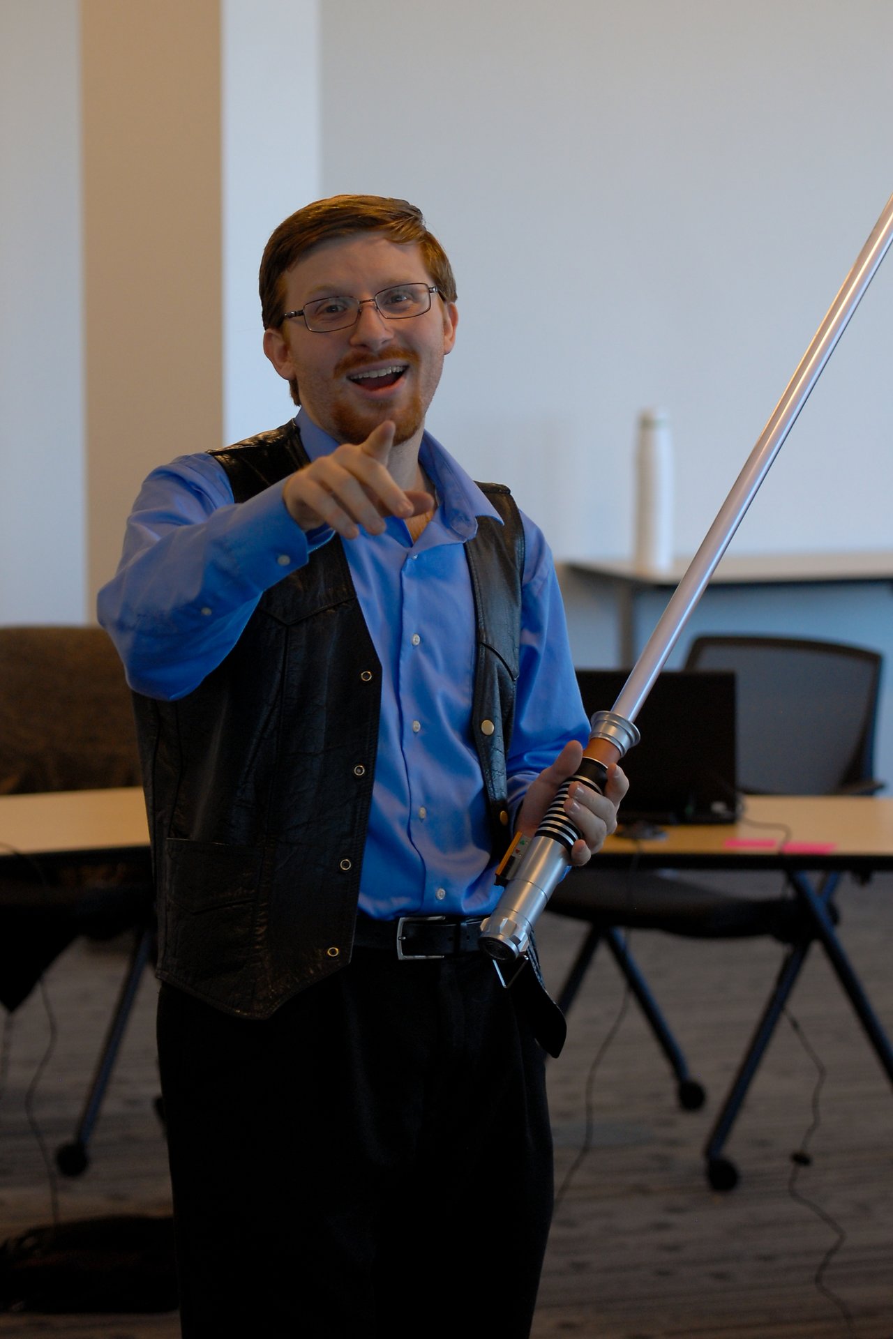 A person in a blue shirt and vest holds a toy lightsaber and points forward with a smile.