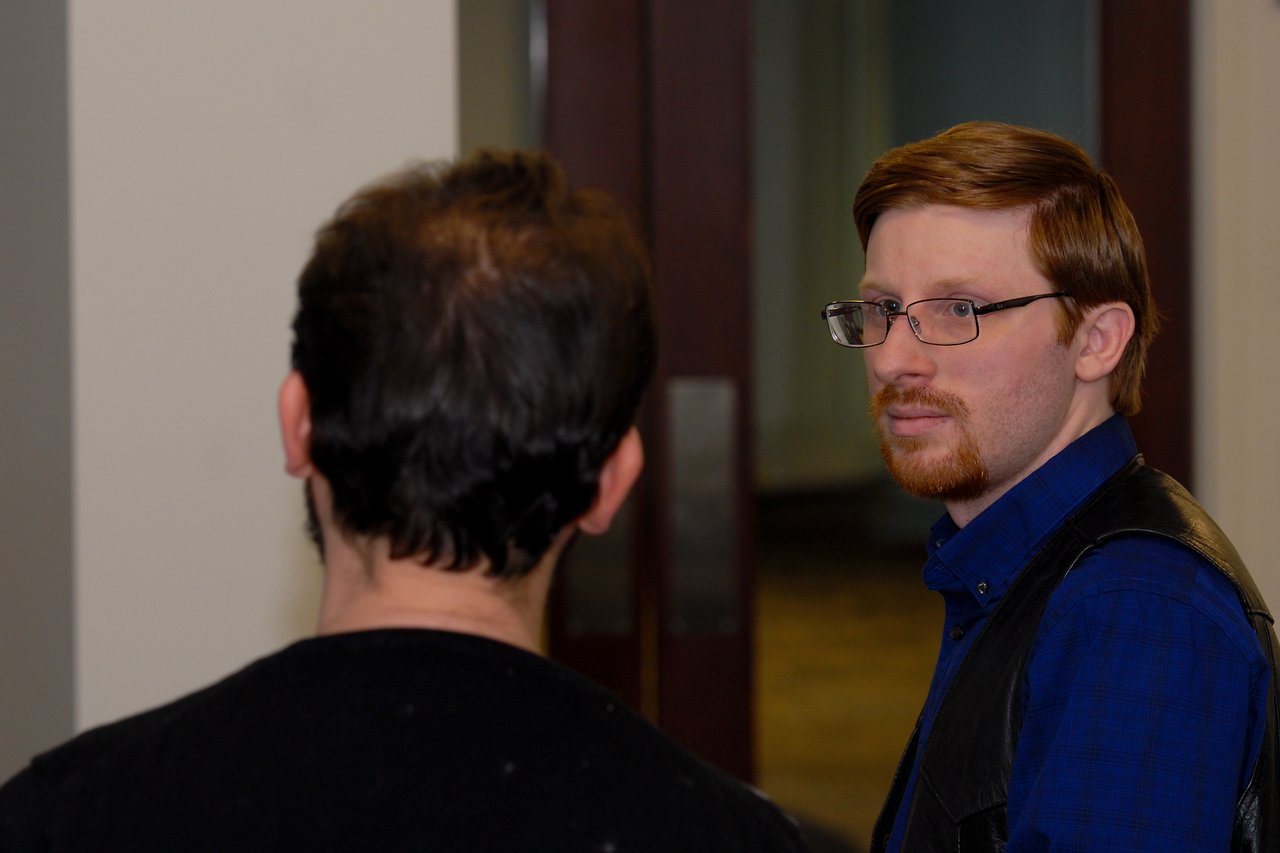 Two people in conversation indoors, one facing away from the camera, the other looking towards them with a serious expression.