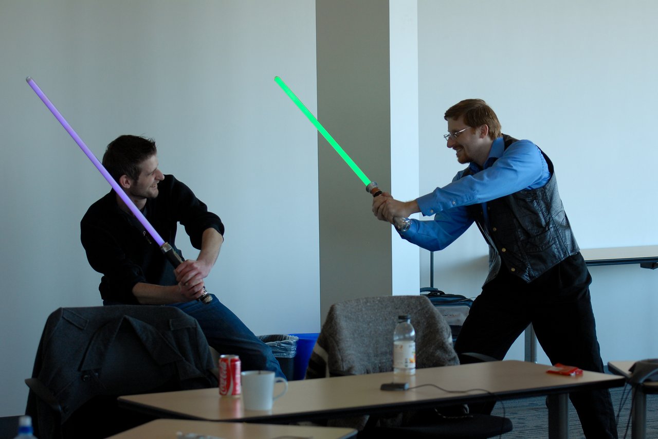 Two people in an office playfully duel with toy lightsabers, one holding a green blade and the other purple.