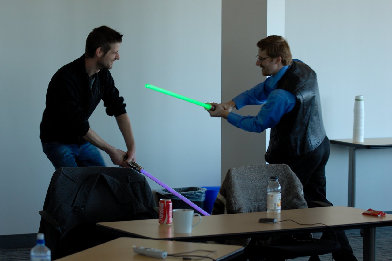 Two people playfully duel with toy lightsabers in an office setting, smiling and engaged in the activity.
