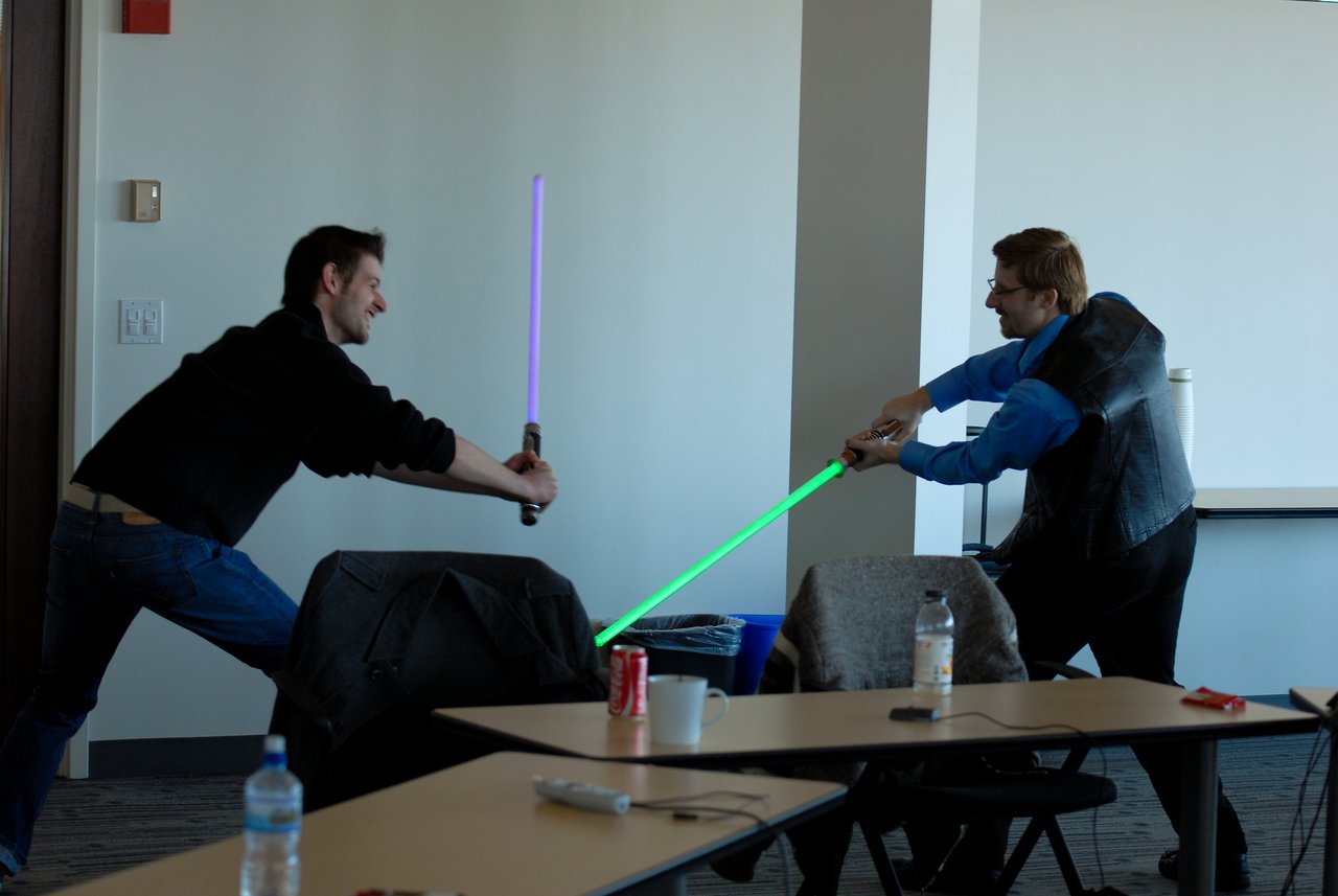 Two people playfully duel with toy lightsabers in an office setting, leaning forward and engaging in the action.