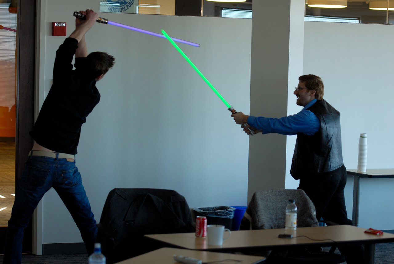 Two people in an office playfully duel with toy lightsabers, one swinging downward and the other blocking.