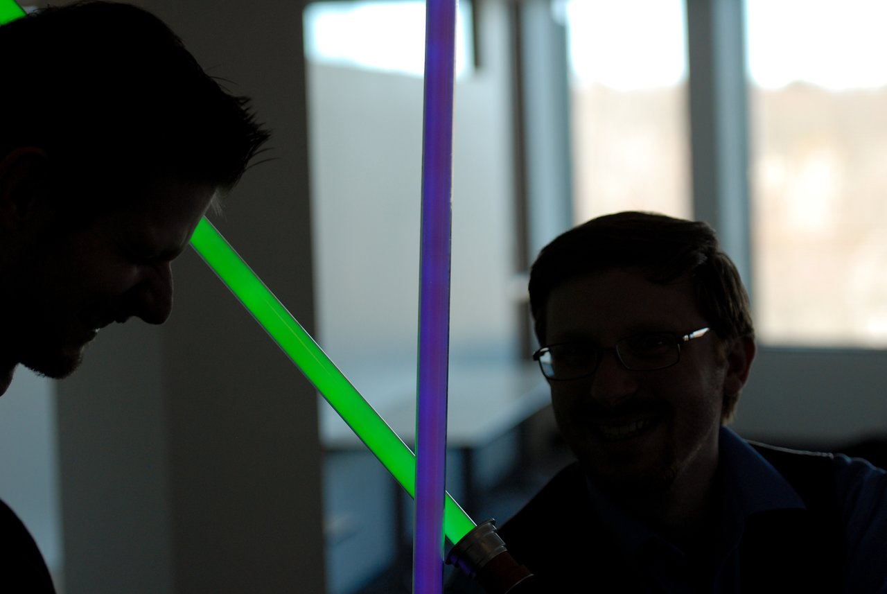 Two people smiling while playfully clashing toy lightsabers, one green and one purple, in an indoor setting.