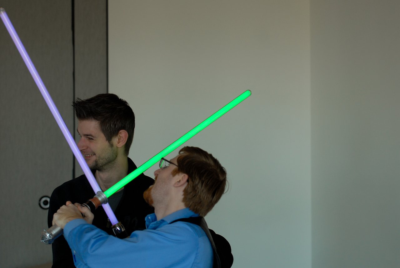 Two people playfully duel with toy lightsabers, one green and one purple, while smiling indoors.