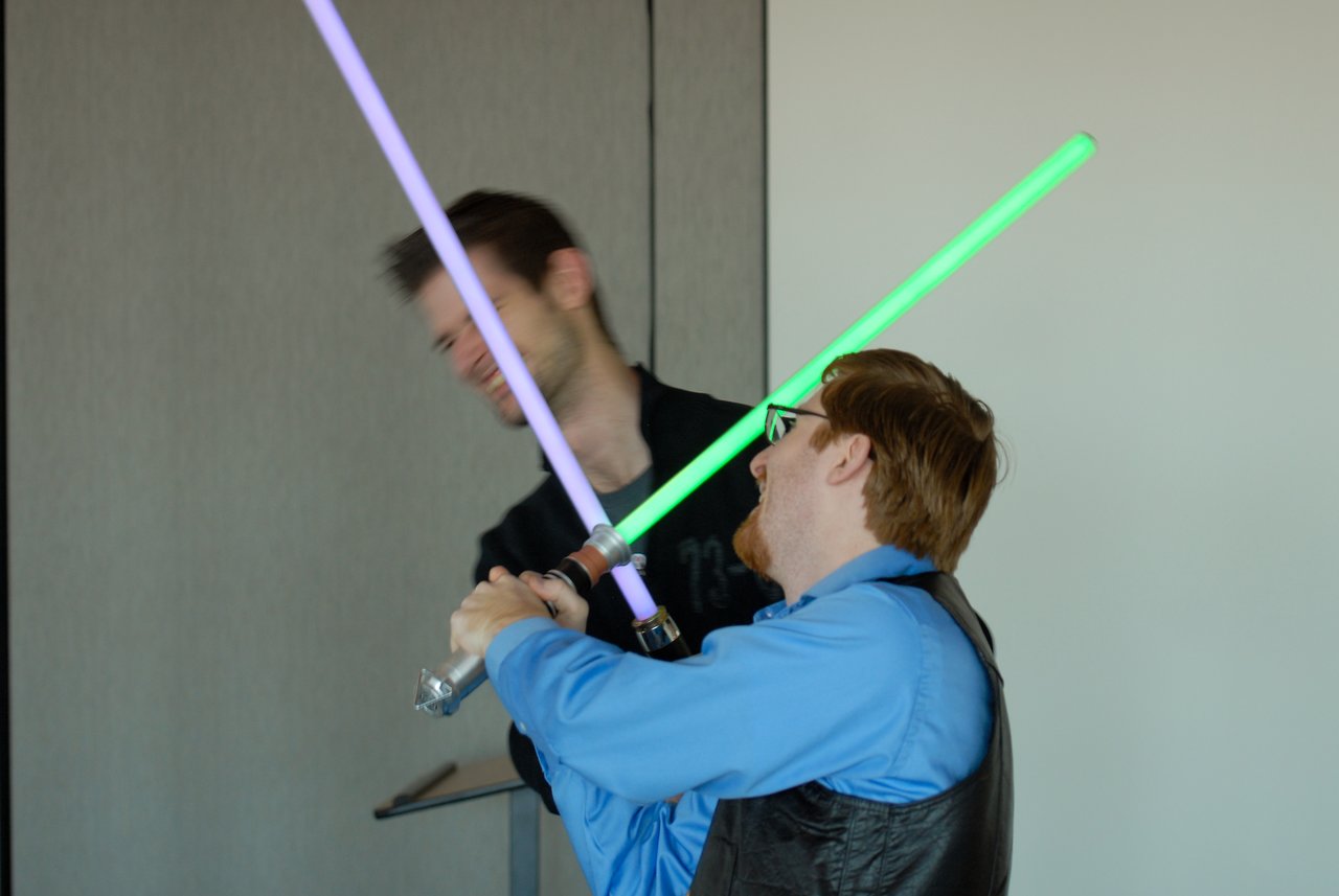 Two people playfully duel with toy lightsabers, one with a green blade and the other with a purple blade.