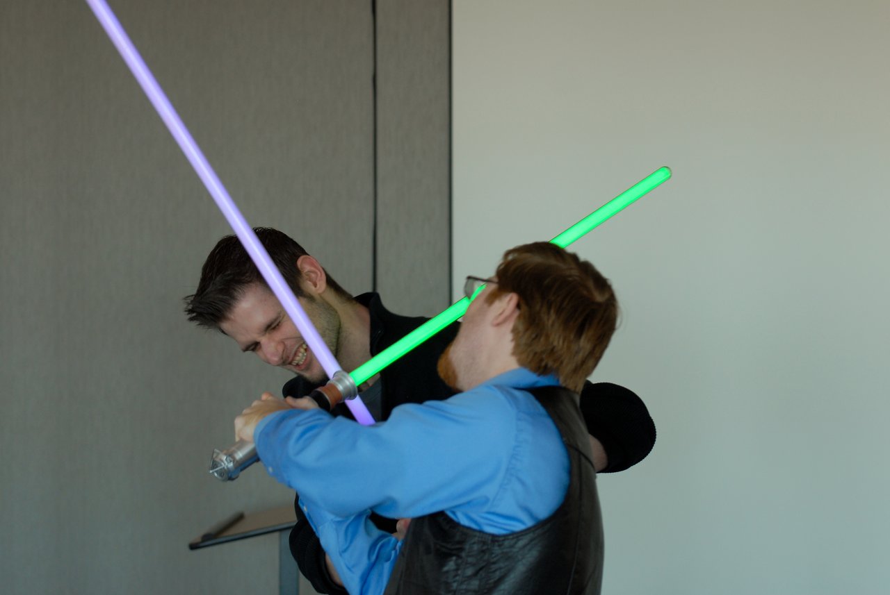 Two people playfully duel with toy lightsabers, one in green and the other in purple, smiling during the interaction.