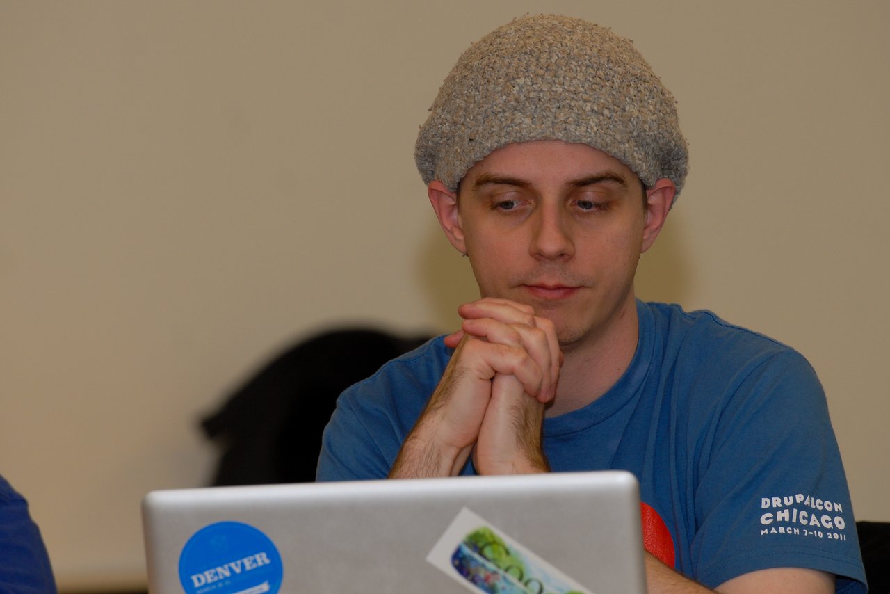 A person in a blue shirt and knit hat looks at a laptop with hands clasped together.
