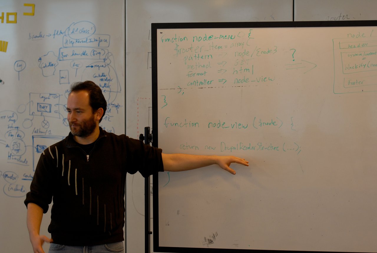 A person gestures toward a whiteboard filled with handwritten code and diagrams during a presentation or discussion.