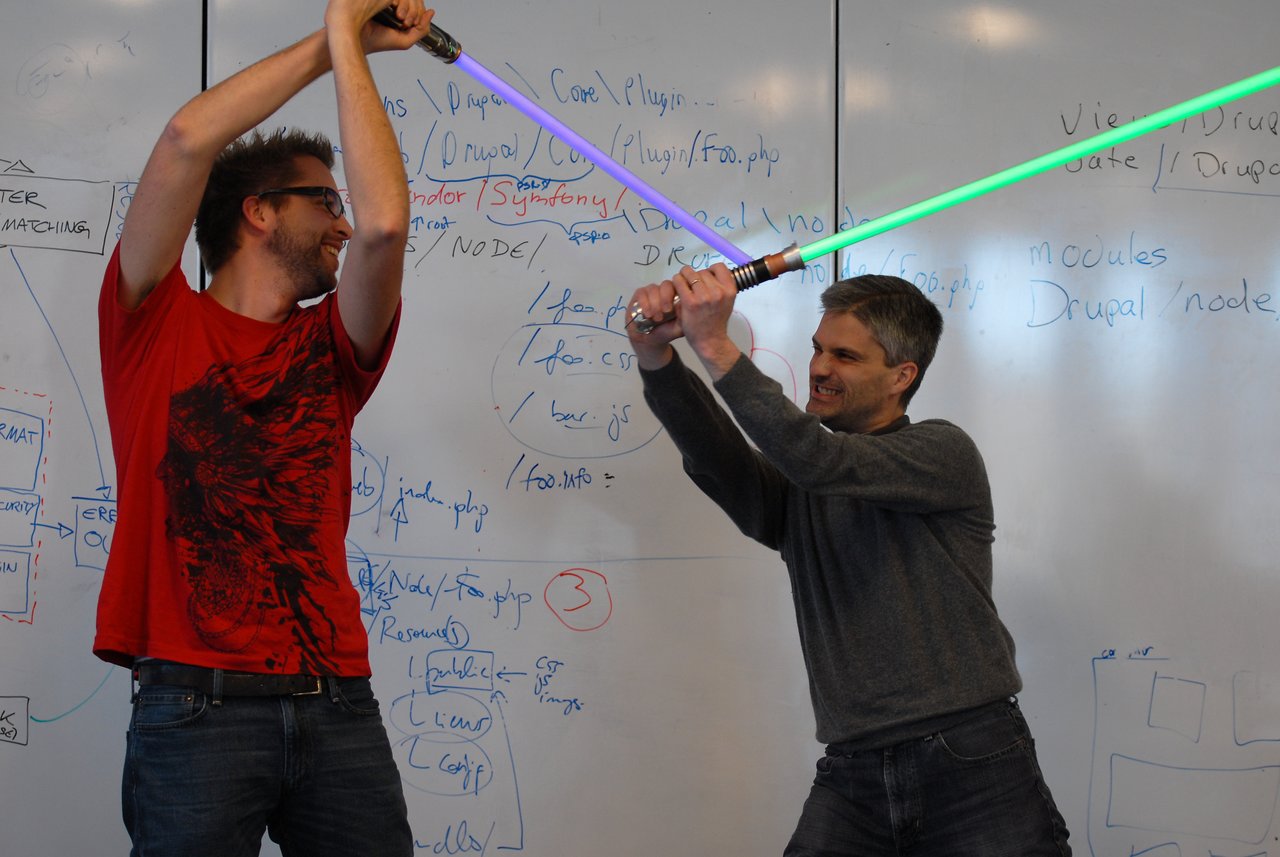 Two people smiling and playfully dueling with toy lightsabers in front of a whiteboard filled with notes.